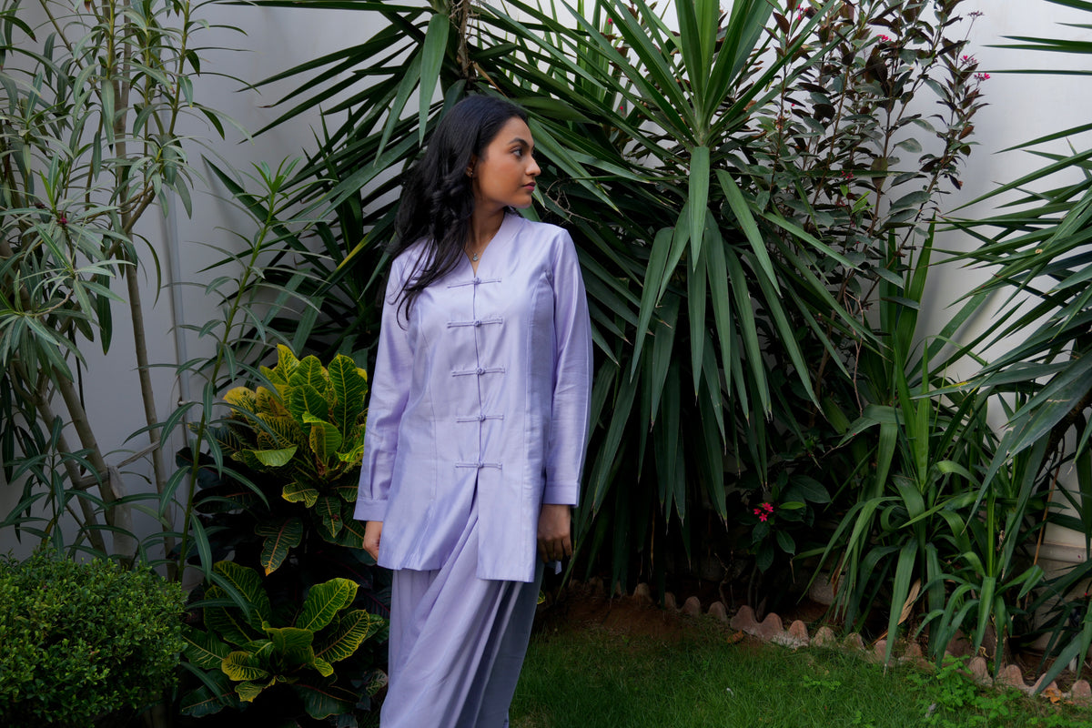 Lilac theory two-piece set featuring mandarin top and matching voluminous balloon pants stands in a lush garden with green plants and a white wall in the background. alisho by alishba kashif